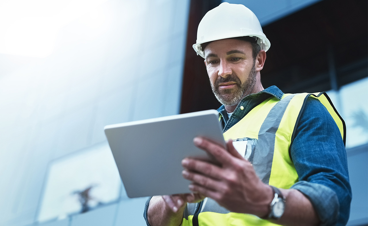 Byggnadsarbetare i hjälm och reflexväst använder en surfplatta utomhus framför en byggnad.